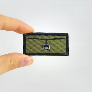 May include: Olive green rectangular patch with a black border. The patch features an embroidered design of a cable car suspended from a cable. The cable car is white with black details. The patch is held by a hand.