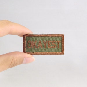 May include: A green and brown embroidered patch with the word "OKAYEST" in brown thread.