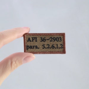May include: A rectangular embroidered patch with a tan background and a brown border. The patch features black embroidered text that reads AFI 36-2903 and para. 5.2.6.12, arranged in two lines across the center of the fabric surface.