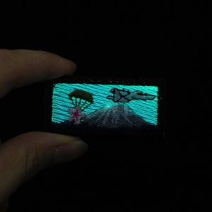 May include: A glow-in-the-dark embroidered patch featuring a mountain, a parachute, and a plane. The mountain is white with a green tree on top. The parachute is pink and the plane is gray. The background is a blue and green gradient.