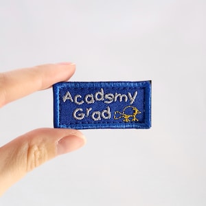 May include: A blue embroidered patch with the words "Academy Grad" in white lettering. A yellow bird is embroidered on the right side. The patch has a blue border and is rectangular in shape.
