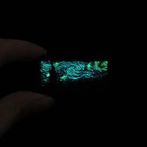 Puede incluir: Un pequeño objeto rectangular brilla con una representación de la pintura "La noche estrellada" de Van Gogh. La obra presenta pinceladas azules y verdes arremolinadas, con una silueta oscura de un árbol y estrellas.