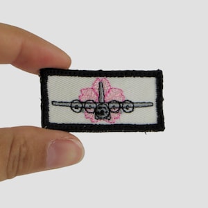 May include: A white embroidered patch with a black border. The patch features a gray airplane with pink flower petals around it.
