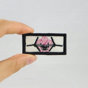 Op de afbeelding: Geborduurde patch met een roze en zwart ontwerp van een gestileerd vliegtuig. De patch heeft een witte achtergrond en een zwarte rand. Het ontwerp is gedetailleerd en gecentreerd, geschikt voor kleding of accessoires.