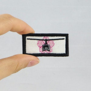 May include: A black and white embroidered patch with a helicopter inside a pink flower. The patch has a black border and is on a white background.