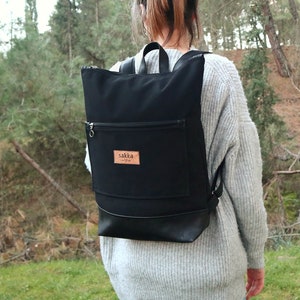 Pode incluir: Uma mochila de lona preta com fundo de couro e um remendo de couro marrom com a palavra "Sakka" nele. A mochila está sendo usada por uma pessoa em um ambiente florestal.