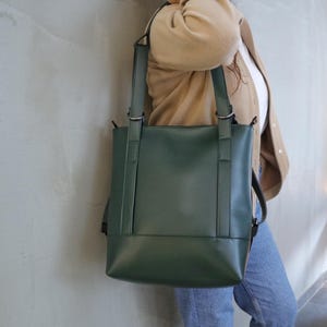 May include: A dark green tote bag with a rectangular shape and two long straps. The bag has a minimalist design and is made of a smooth material. The straps are attached to the bag with silver metal rings. The bag is being carried over the shoulder.