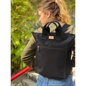 Könnte beinhalten: Eine schwarze Canvas-Tragetasche mit einem oberen Griff und verstellbaren Trägern. Die Tasche hat eine Reißverschlusstasche auf der Vorderseite und ein kleines Leder-Patch mit dem Wort "sakka" darauf.