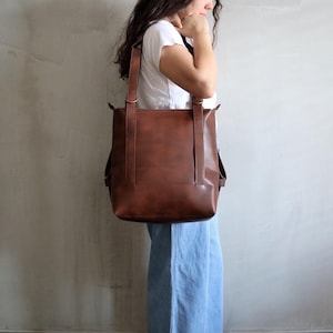 Pode incluir: Uma bolsa tote grande e retangular na cor marrom com alças verticais e anéis de metal no topo. A bolsa apresenta um acabamento liso e marrom sólido, sendo usada sobre o ombro. Possui um design estruturado com fixações nas laterais.
