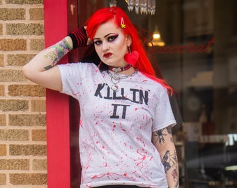 May include: A person wearing a white t-shirt with the text "KILLIN IT" and red splatter design, paired with a black and red plaid skirt, and purple platform boots. The outfit includes black and red striped arm warmers and knee-high socks.