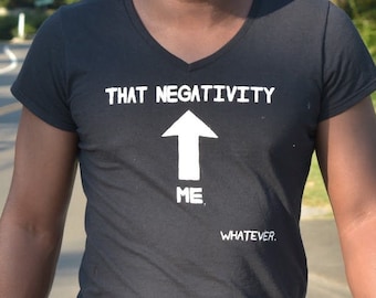 May include: Black V-neck t-shirt with the text "THAT NEGATIVITY ME WHATEVER" and an arrow graphic. The person is wearing sunglasses and blue jeans. The background includes trees and a crosswalk.