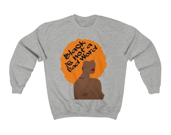 May include: A light gray sweatshirt featuring a graphic of a woman with orange hair and the text "Black is not a bad word." The woman has red lips and a tattoo on her upper arm. The sweatshirt is a casual style.
