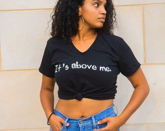 May include: Black V-neck t-shirt with the text "It's above me." The shirt is knotted at the waist and paired with blue jeans. The model is wearing gold hoop earrings and a black bracelet.