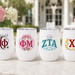 Puede incluir: Cuatro vasos blancos sin tallo dispuestos en fila. Cada vaso presenta un diseño único de letras griegas: el primero tiene letras granates con un lazo plateado e hiedra, el segundo letras rosas con un lazo y una rosa, el tercero letras azul cerceta con una corona y el cuarto letras rojas con un búho dorado y una rama de laurel.
