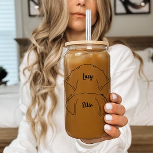 May include: A clear glass tumbler with a bamboo lid and a straw. The tumbler contains a brown liquid and features two dog silhouettes with the names "Lucy" and "Ellie" printed on it.