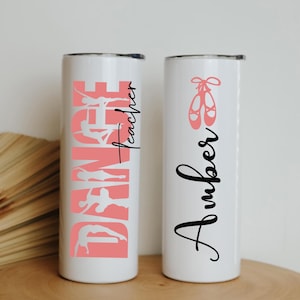 Pode incluir: Dois copos brancos de aço inoxidável com designs rosa e preto. Um copo tem a palavra "Dance" em rosa com uma silhueta de uma bailarina e as palavras "Teacher" em preto. O outro copo tem a palavra "Amber" em preto com um par de sapatilhas de balé rosa.
