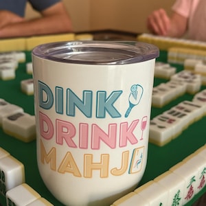 May include: White tumbler with the words "DINK DRINK MAHJI" in teal, pink, and yellow. A cartoon illustration of a paddle and a wine glass is also on the tumbler. Mahjong tiles are on a green surface.