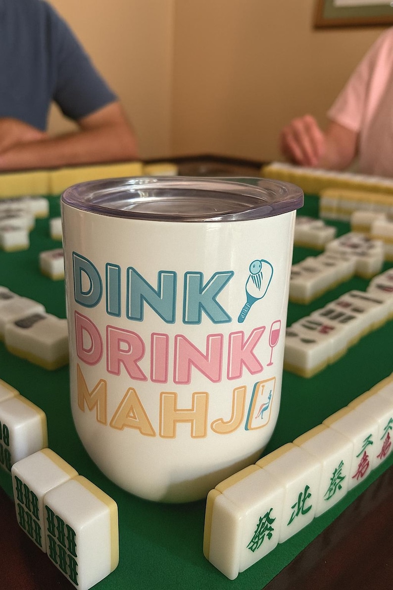 May include: White tumbler with the words "DINK DRINK MAHJI" in teal, pink, and yellow. A cartoon illustration of a paddle and a wine glass is also on the tumbler. Mahjong tiles are on a green surface.