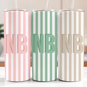 May include: Three stainless steel tumblers with vertical striped patterns in pink, green, and beige. Each tumbler features the initials "NB" in a matching solid color. The tumblers have a straw and are designed for cold beverages.