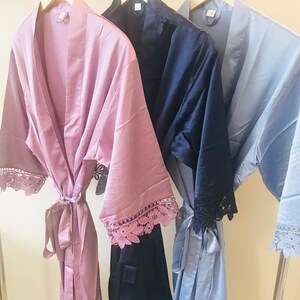 May include: Three satin robes with lace trim hanging on wooden hangers. The robes are in dusty rose, navy blue, and light blue.