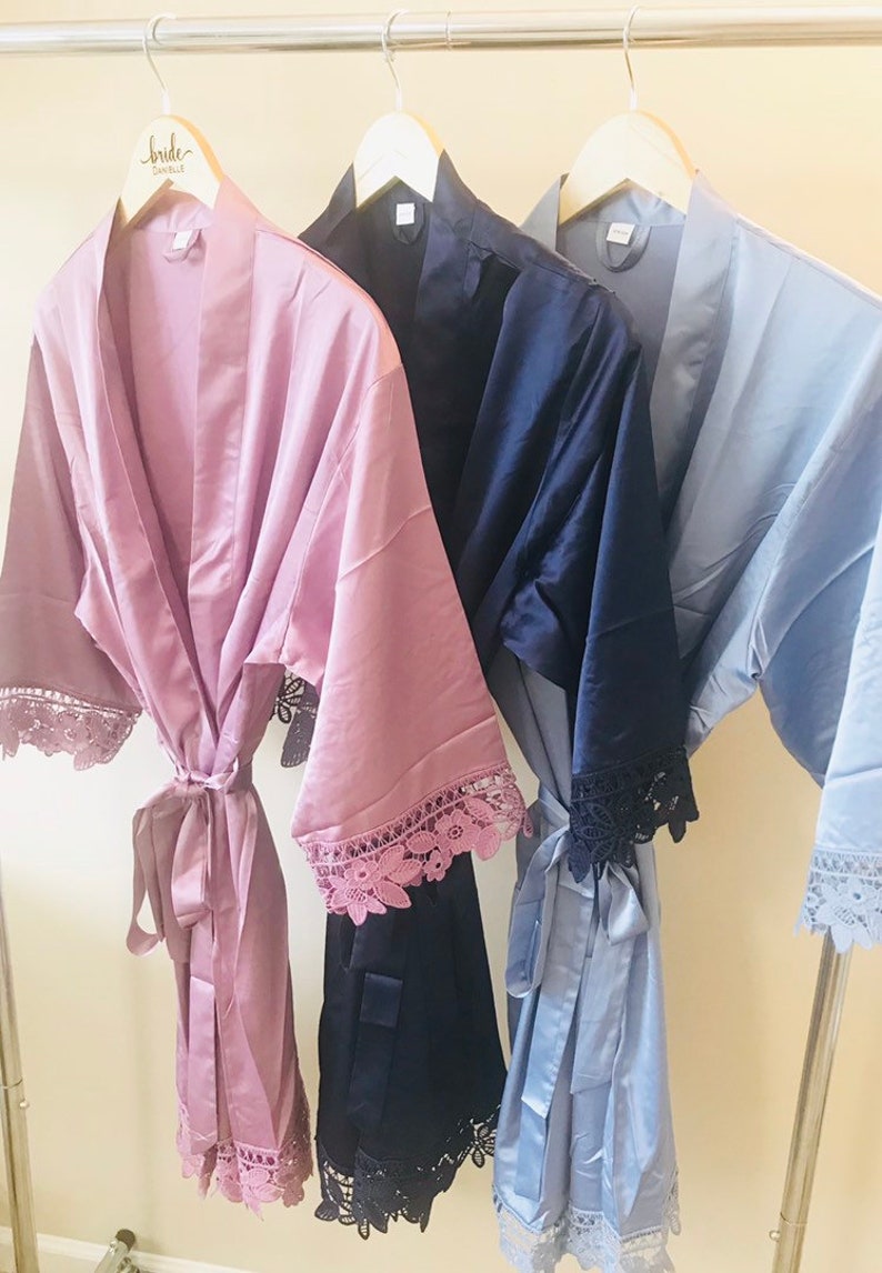 May include: Three satin robes with lace trim hanging on wooden hangers. The robes are in dusty rose, navy blue, and light blue.