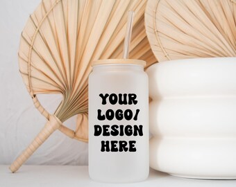 Pode incluir: Um copo de vidro branco fosco com tampa de bambu e um canudo preto. O copo tem o texto "YOUR LOGO/DESIGN HERE" impresso na frente.