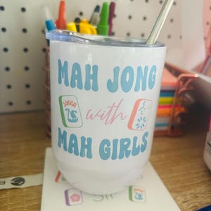 May include: White travel mug with a blue and pink design that says "Mah Jong with Mah Girls". The mug has a straw and is sitting on a white coaster with mah jong tiles.