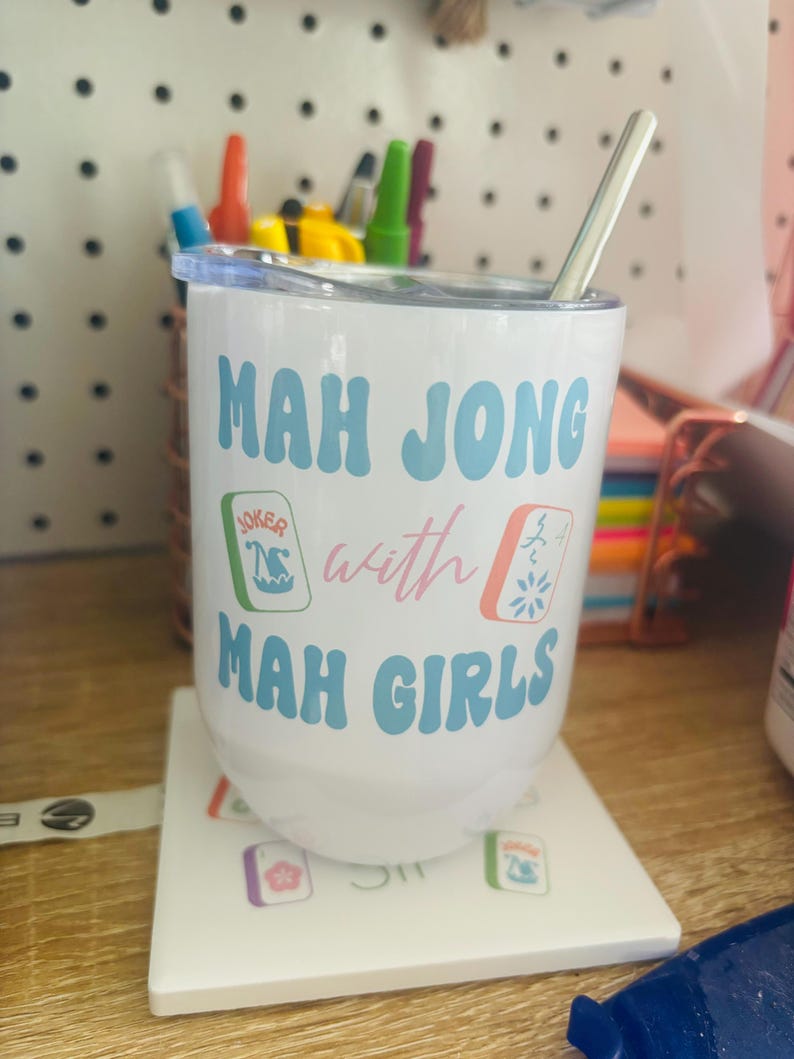 May include: White travel mug with a blue and pink design that says "Mah Jong with Mah Girls". The mug has a straw and is sitting on a white coaster with mah jong tiles.