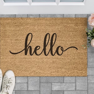 Puede incluir: Una alfombra de coco marr&oacute;n con la palabra "hello" escrita en cursiva negra.