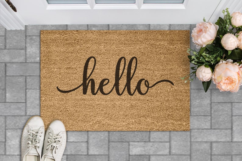 Puede incluir: Una alfombra de coco marr&oacute;n con la palabra "hello" escrita en cursiva negra.