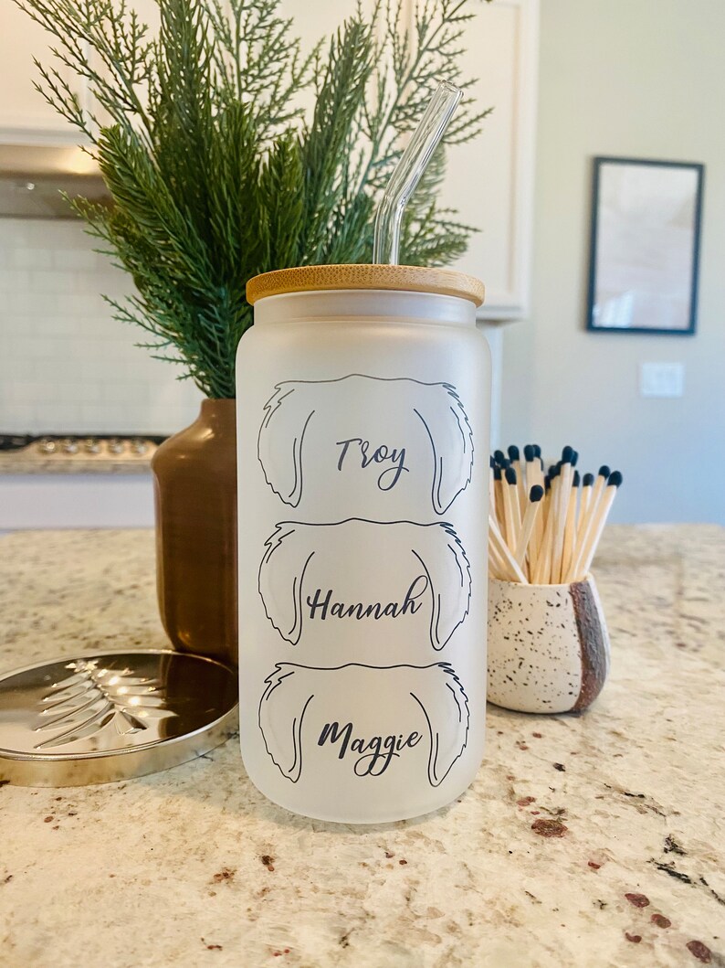May include: A frosted glass water bottle with a bamboo lid and a clear reusable straw. The bottle has three dog silhouettes with the names "Troy", "Hannah", and "Maggie" printed on them.