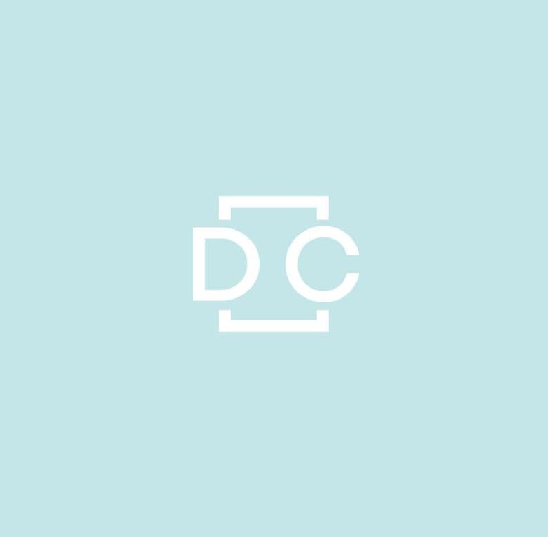 May include: White logo on a light blue background. The logo features the letters "D" and "C" in a sans-serif font, enclosed within a rectangular frame. The design is simple and modern, with a minimalist aesthetic.