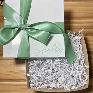 May include: A white gift box with a green satin ribbon and bow. The box is personalized with the name "Shelby". The box is open and filled with white shredded paper.