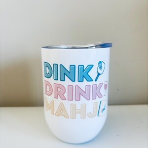 May include: White stemless wine tumbler with a blue lid. The tumbler features the words "DINK! DRINK! MAHJ!" in blue, pink, and beige lettering, along with pickleball and wine glass graphics.