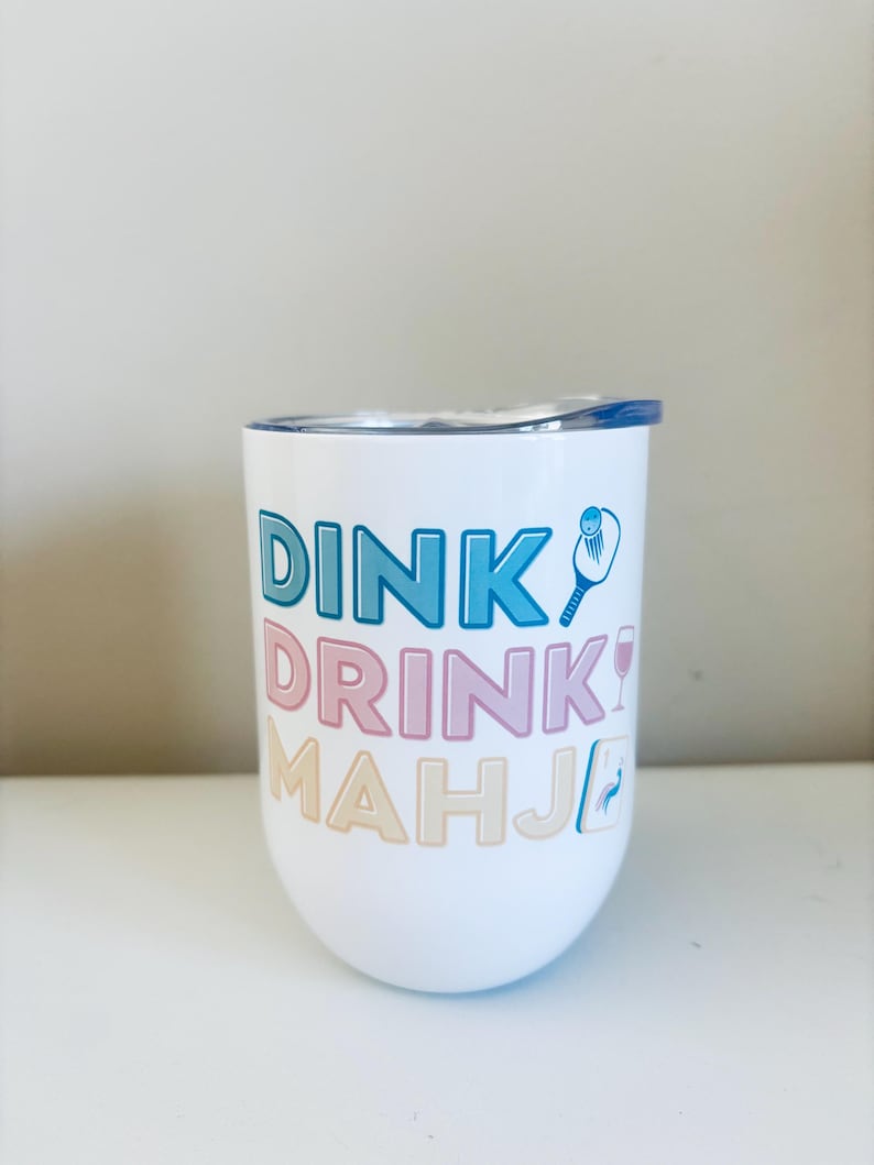 May include: White stemless wine tumbler with a blue lid. The tumbler features the words "DINK! DRINK! MAHJ!" in blue, pink, and beige lettering, along with pickleball and wine glass graphics.