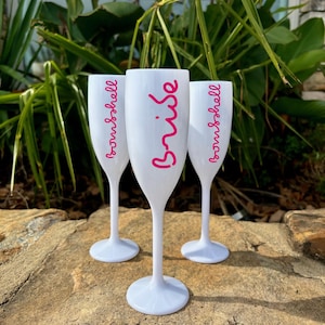 Op de afbeelding: Drie witte plastic champagnefluiten met roze opschrift. De middelste fluit zegt "Bride" en de andere twee zeggen "Bridesmaid".