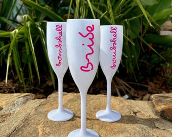 Può includere: Tre flute da champagne in plastica bianca con scritte rosa. La flute centrale dice "Bride" e le altre due dicono "Bridesmaid".