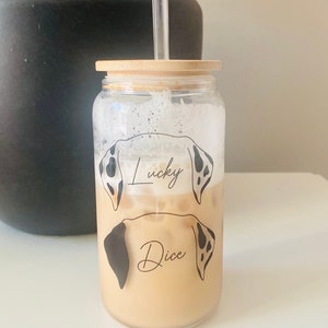 May include: A clear glass jar with a bamboo lid and a straw. The jar features a design of two dalmatian dog heads with the names "Lucky" and "Dice" written on them. The jar is filled with a light brown liquid.