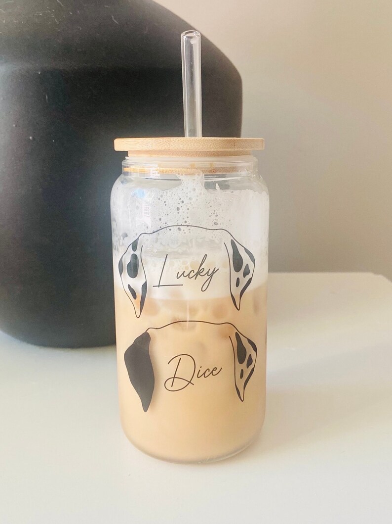 May include: A clear glass jar with a bamboo lid and a straw. The jar features a design of two dalmatian dog heads with the names "Lucky" and "Dice" written on them. The jar is filled with a light brown liquid.