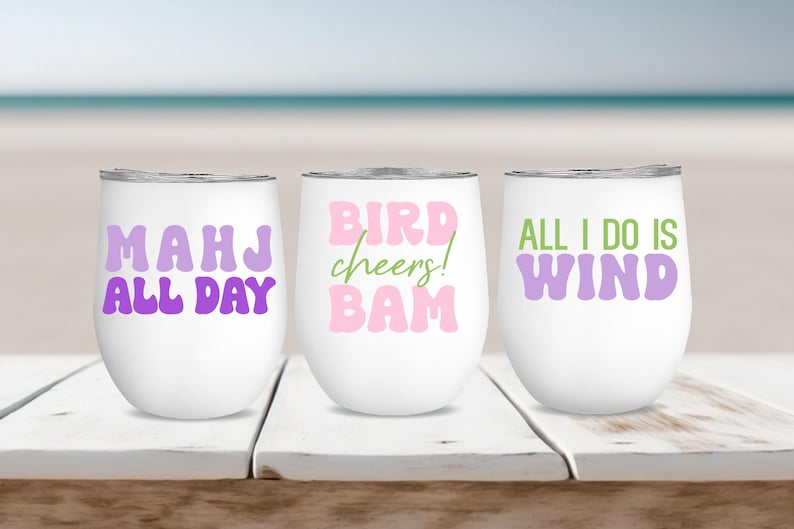 May include: Three white stainless steel wine tumblers with different phrases printed on them. The first tumbler says "Mahj all day" in purple letters. The second tumbler says "Bird cheers! Bam" in pink letters. The third tumbler says "All I do is wind" in purple letters.