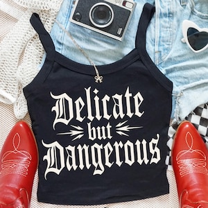 Könnte beinhalten: Ein Mode-Flatlay mit einem schwarzen Tanktop mit dem Schriftzug "Delicate but Dangerous", Jeansshorts, herzförmigen Sonnenbrillen und roten Cowboy-Stiefeln. Eine kleine Kamera und ein karierter Schal sind ebenfalls enthalten.