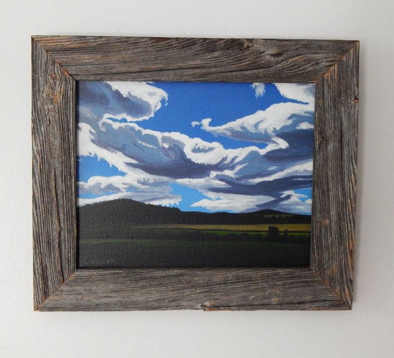 Puede incluir: Una pintura enmarcada de un paisaje con un cielo azul y nubes blancas. La pintura est&aacute; en un marco de madera r&uacute;stico.