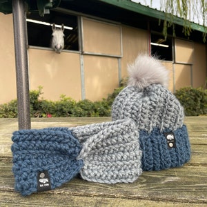 以下が含まれることがあります： 手作りのかぎ針編み冬物アクセサリーセット。青とグレーのヘッドバンドと、グレーのフェイクファーポンポンが付いたお揃いの帽子が含まれます。各アイテムには、ロゴと「Est. 1996」の文字が入った小さな黒いタグが付いています。