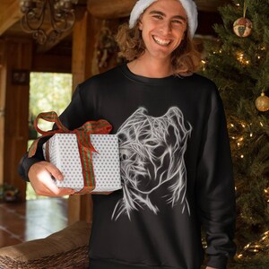 Op de afbeelding: Een zwart sweatshirt met een witte afbeelding van een gestileerd hondenhoofd. De persoon die het sweatshirt draagt, glimlacht en houdt een ingepakt cadeau vast.