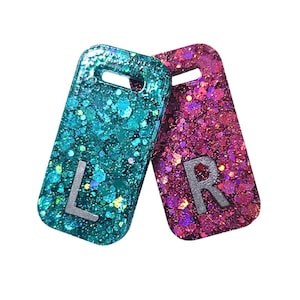Pode incluir: Duas etiquetas de resina brilhantes, uma turquesa e outra magenta, com letras prateadas "L" e "R". Cada etiqueta tem formato retangular com um pequeno orifício na parte superior. As etiquetas são cobertas com glitter brilhante.