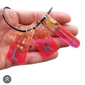 Peut inclure: Un collier fait à la main avec trois pendentifs rectangulaires. Les pendentifs sont un dégradé de rose et de rouge, avec des paillettes dorées et des lettres argentées ou un motif de flèche. Le collier a un cordon noir.