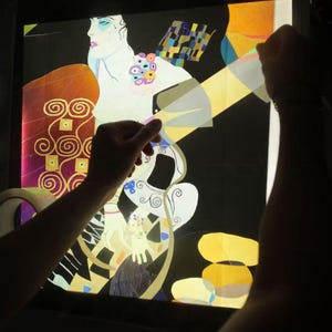 May include: A person is holding a light box with a colorful abstract design. The design features a woman with a flowing dress and a geometric pattern.