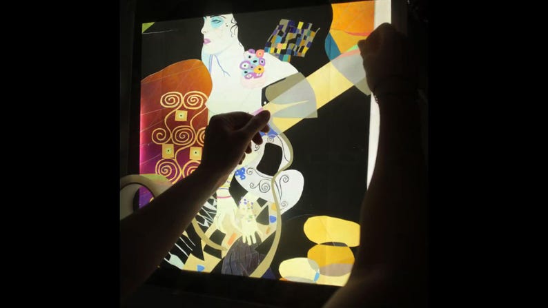 May include: A person is holding a light box with a colorful abstract design. The design features a woman with a flowing dress and a geometric pattern.