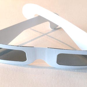 May include: White cardboard 3D glasses with gray lenses. The glasses are folded in half, with the lenses facing the viewer.