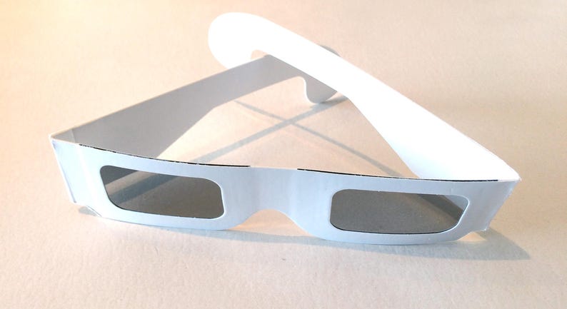 May include: White cardboard 3D glasses with gray lenses. The glasses are folded in half, with the lenses facing the viewer.